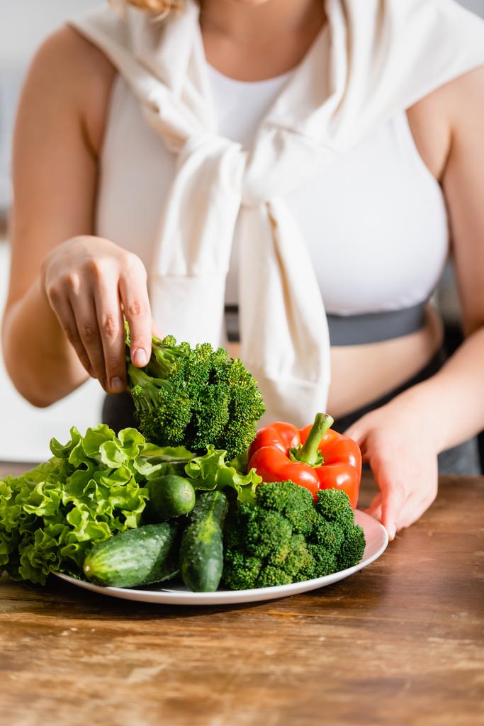 обрезанный вид женщины, держащей свежие брокколи возле спелых овощей на тарелке  - Фото, изображение