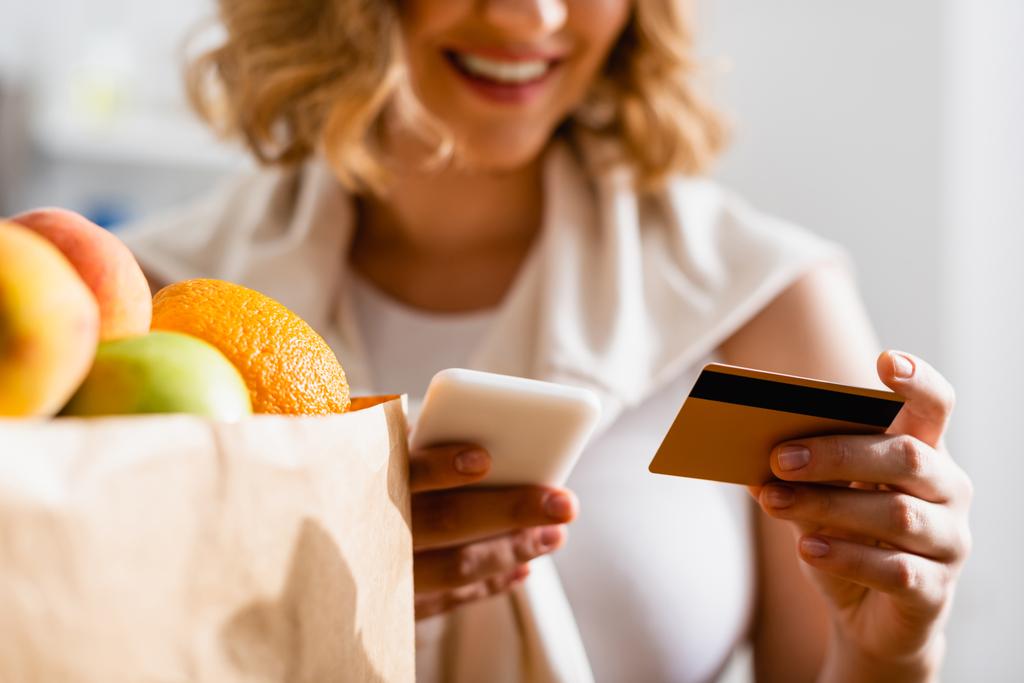 částečný pohled na ženu držící smartphone a kreditní kartu v blízkosti ovoce v papírové tašce  - Fotografie, Obrázek