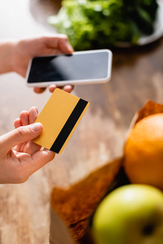 Selektiver Fokus der Frau, die Kreditkarte und Smartphone mit leerem Bildschirm in der Nähe von Früchten in Papiertüte hält  - Foto, Bild