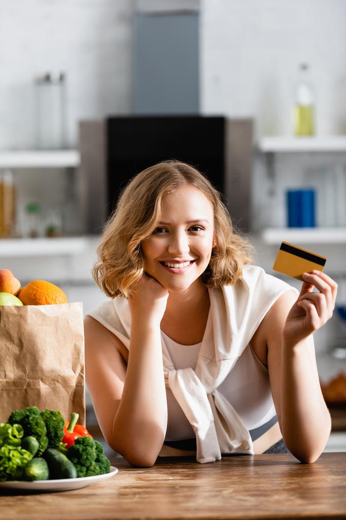 mujer sosteniendo tarjeta de crédito cerca de bolsa de papel y plato con verduras en la mesa  - Foto, imagen