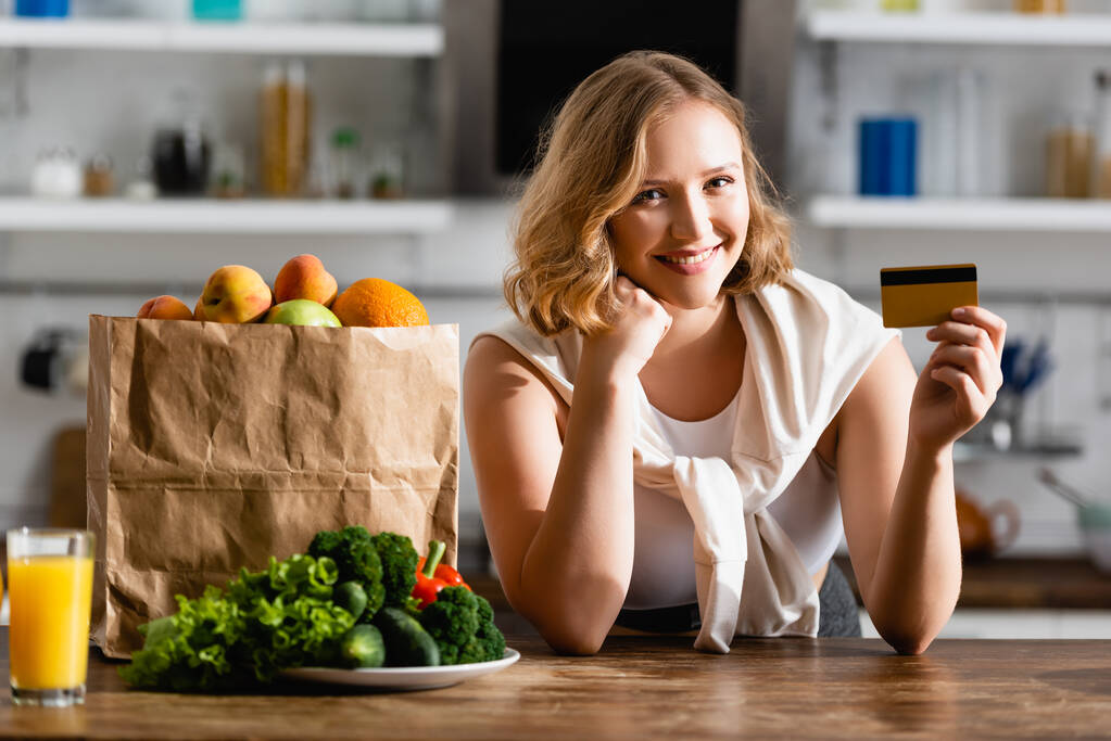 young woman holding credit card near paper bag and plate with vegetables on table  - Photo, Image