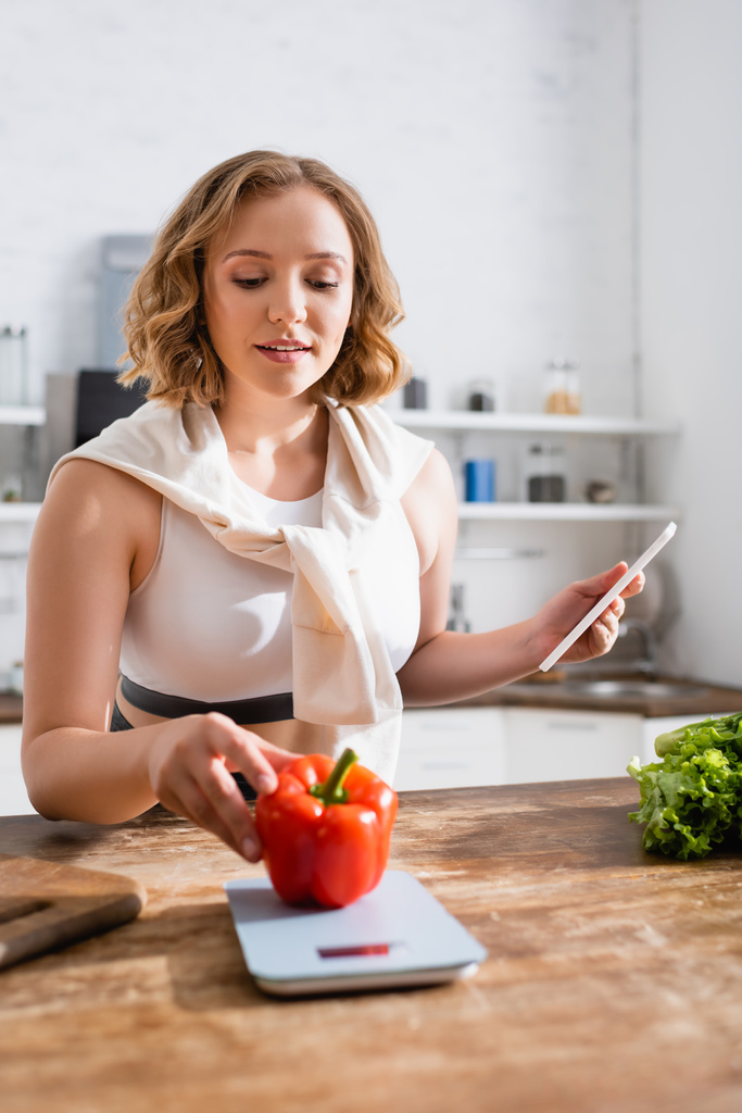 γυναίκα αγγίζοντας bell pepper σε κλίμακες κουζίνας και κρατώντας smartphone  - Φωτογραφία, εικόνα