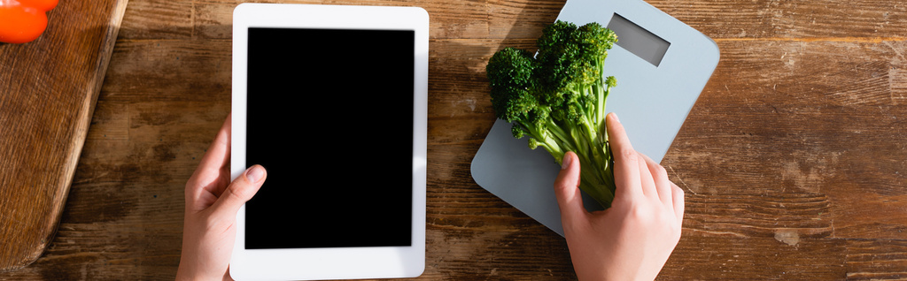 imagem horizontal de mulher segurando brócolis perto de balanças de cozinha e tablet digital com tela preta
  - Foto, Imagem