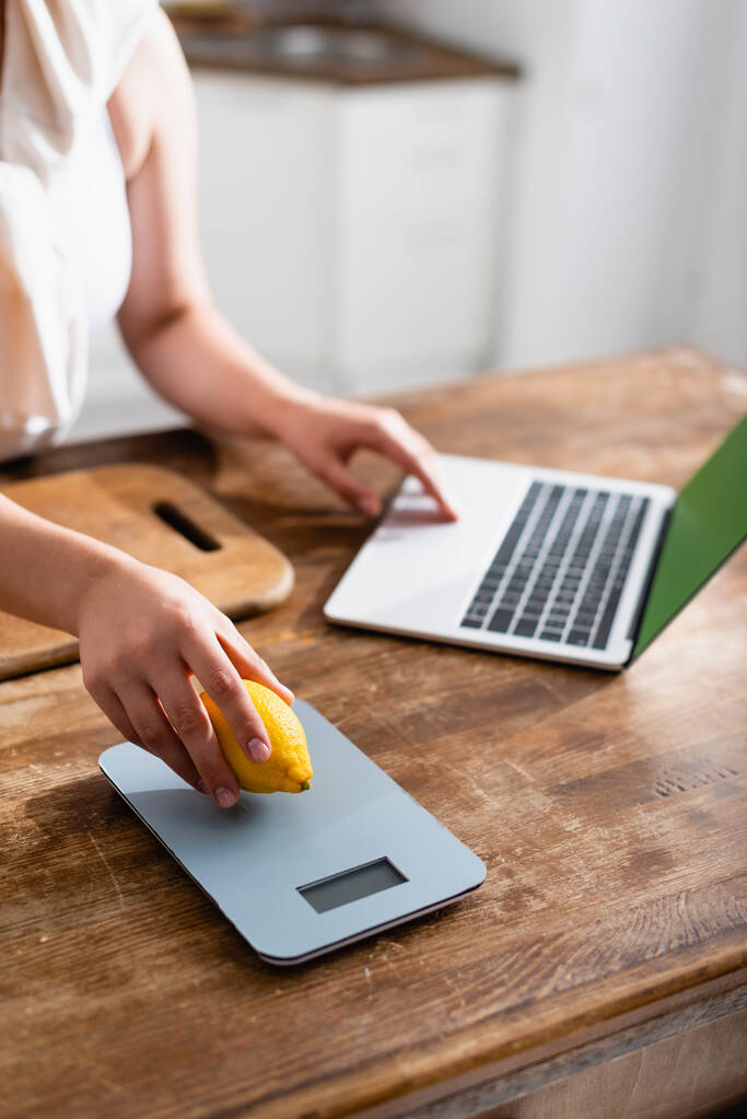 abgeschnittene Ansicht einer Frau, die Zitrone in der Nähe von Küchenwaagen hält und Laptop benutzt  - Foto, Bild