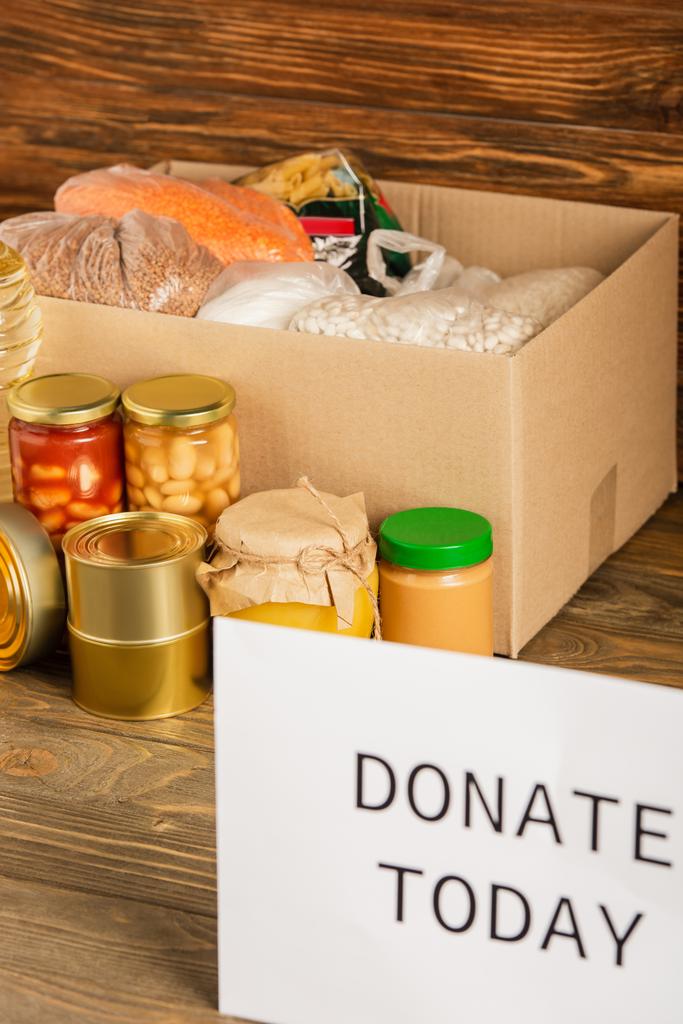 selective focus of cardboard box with food and donate today card on wooden background, charity concept - Photo, Image