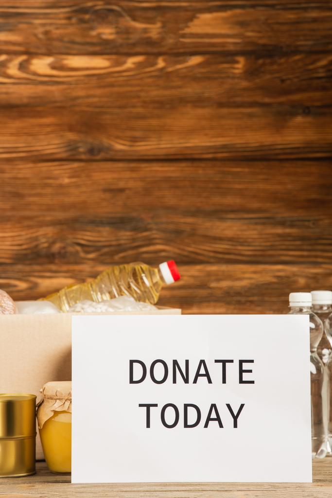 cardboard box with food and donate today card on wooden background, charity concept - Photo, Image