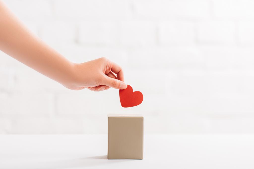 oříznutý pohled na ženskou ruku dávající červené srdce do krabice na bílém pozadí, koncept darování - Fotografie, Obrázek
