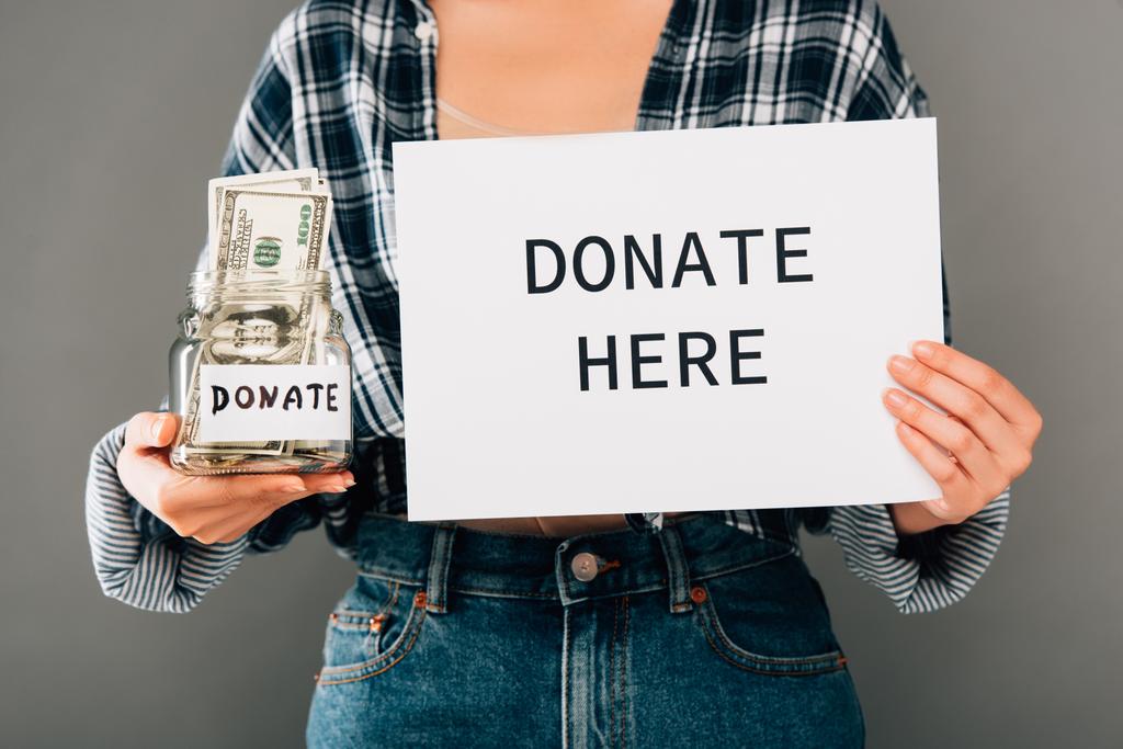 cropped view of woman holding card with donate here lettering and jar with money on grey background - Photo, Image