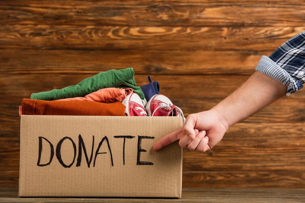 cropped view of man pointing at cardboard box with donate lettering and clothes on wooden background, charity concept - Photo, Image