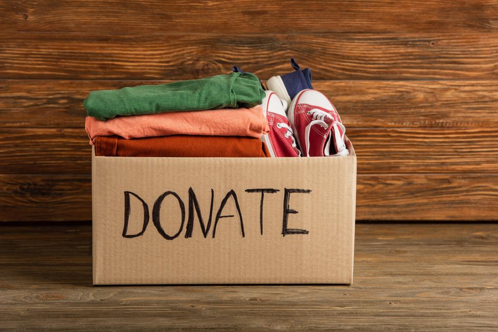 cardboard box with donated clothes and footwear on wooden background, charity concept - Photo, Image