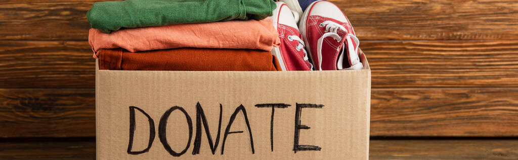 panoramic shot of cardboard box with donated clothes and footwear on wooden background, charity concept - Photo, Image