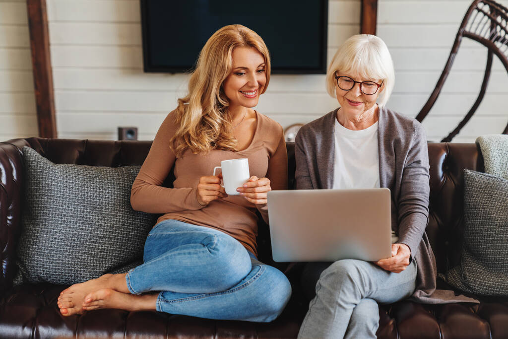 Улыбающаяся пожилая женщина и ее дочь среднего возраста сидят расслабиться на диване и пить кофе в гостиной смотреть видео на ноутбуке вместе - Фото, изображение