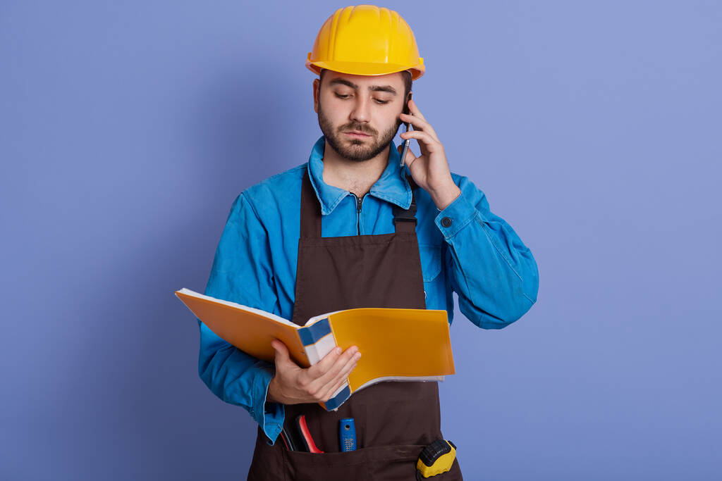 European architect with yellow paper folder in hands reading important information to his partner or bass, while talking via smart phone, wearing helmet and apron, standing against blue wall. - Photo, Image