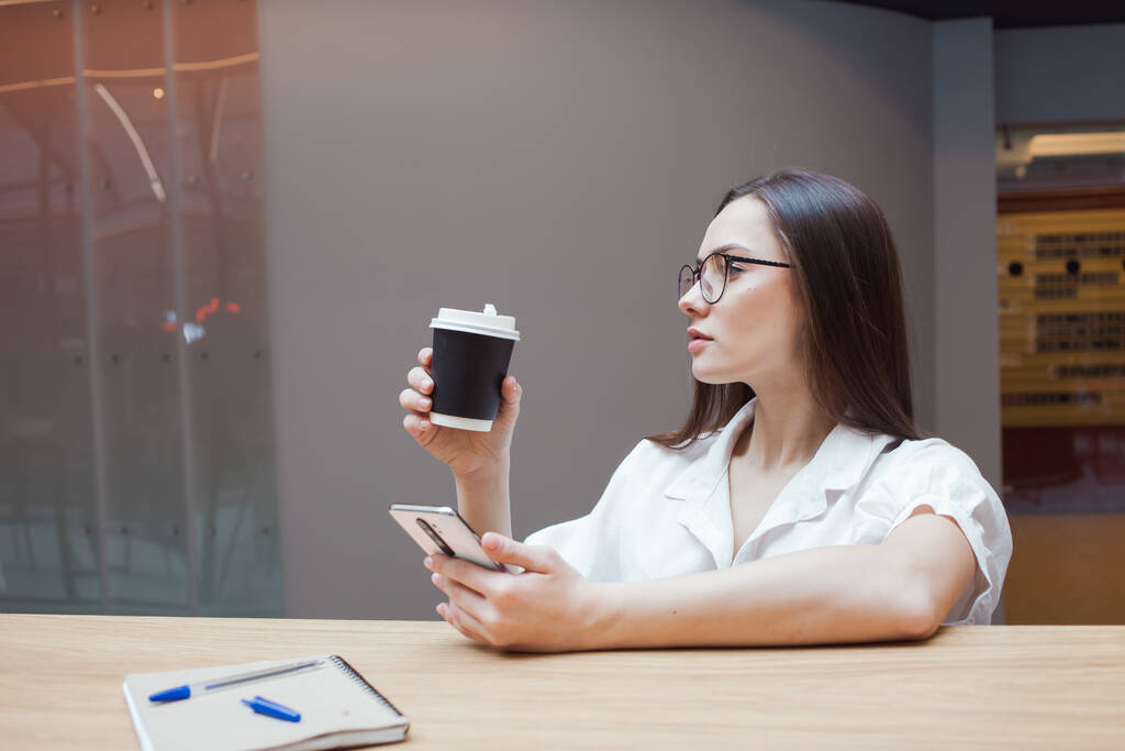 Μαθήτρια κάθεται σε ένα τραπέζι και χρησιμοποιεί μια εφαρμογή στο smartphone της. Μια νεαρή μελαχρινή με γυαλιά και λευκό πουκάμισο. - Φωτογραφία, εικόνα