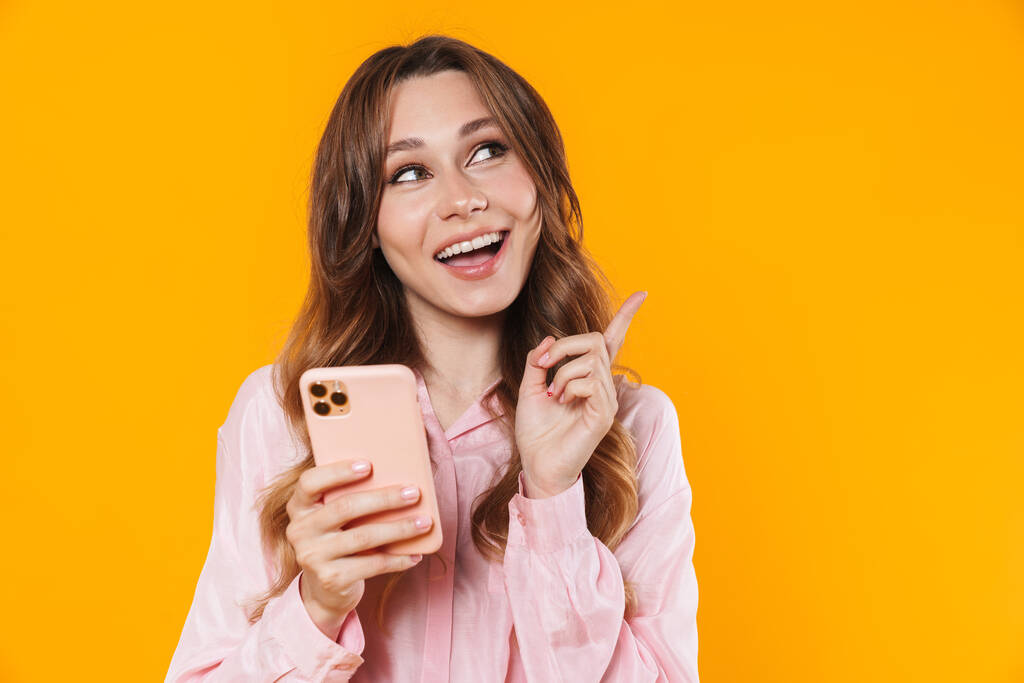 Imagen de una bonita mujer sonriente señalando con el dedo a un lado y usando un teléfono móvil aislado sobre un fondo amarillo - Foto, imagen