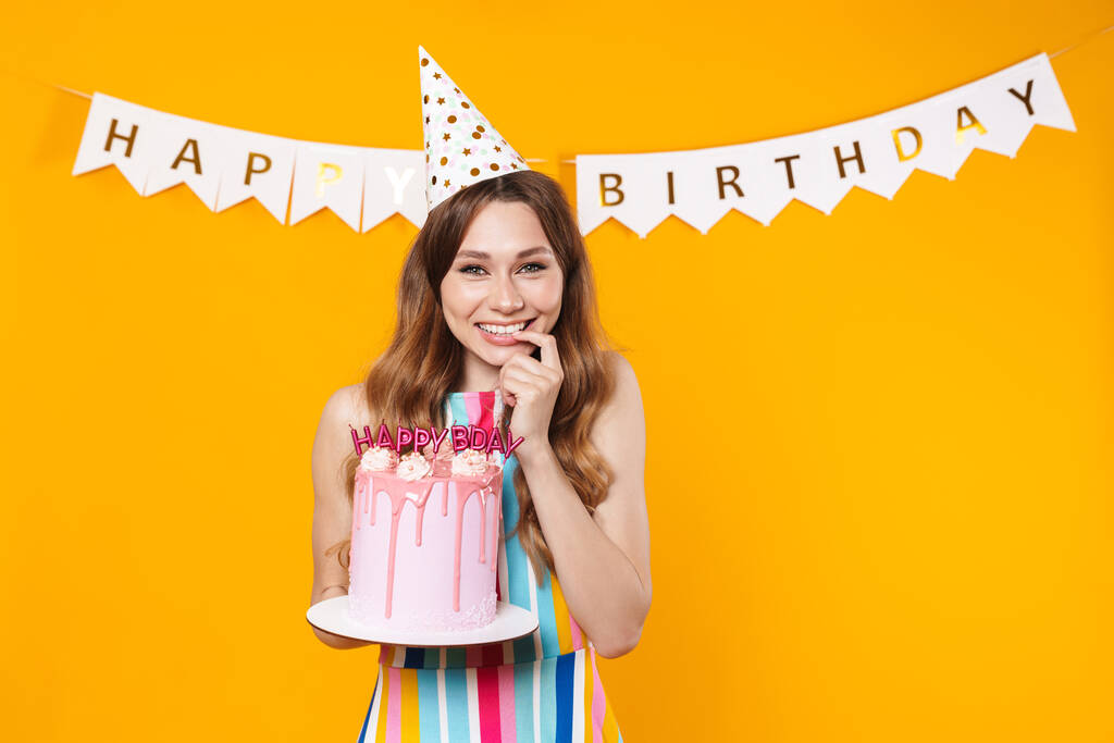 Image de jeune femme excitée dans le cône de fête montrant torte anniversaire et sourire isolé sur fond jaune - Photo, image