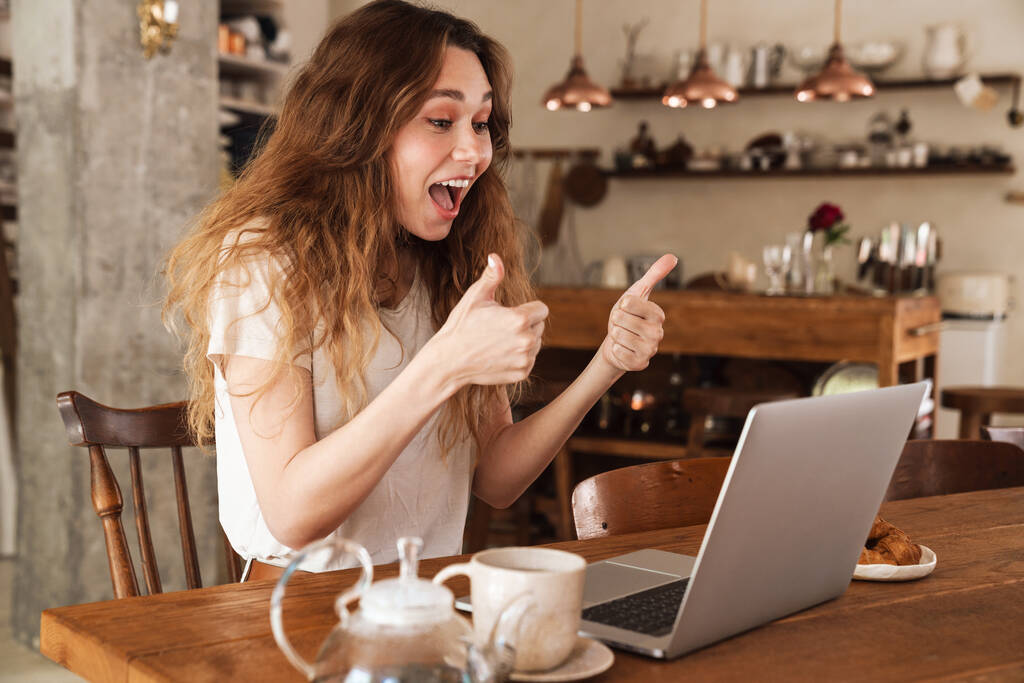 Kaunis nuori hymyilevä tyttö istuu keittiön pöydässä, kannettavan tietokoneen avulla, videopuhelu - Valokuva, kuva