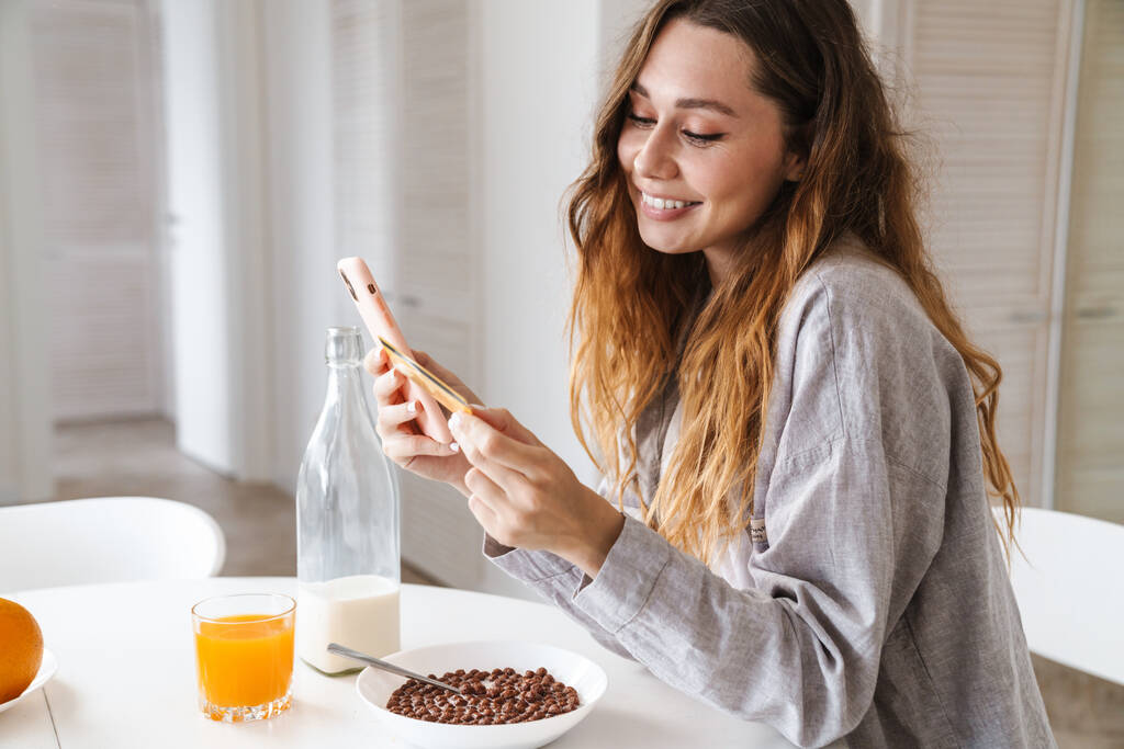 Melko iloinen nuori nainen syö aamiaista istuessaan keittiön pöydässä matkapuhelimen ja luottokortin avulla verkko-ostoksia varten. - Valokuva, kuva
