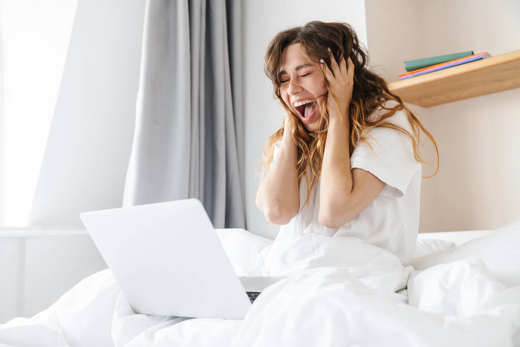 Retrato de mujer excitada jengibre expresando sorpresa y el uso de la computadora portátil mientras está sentado en la cama después del sueño - Foto, imagen