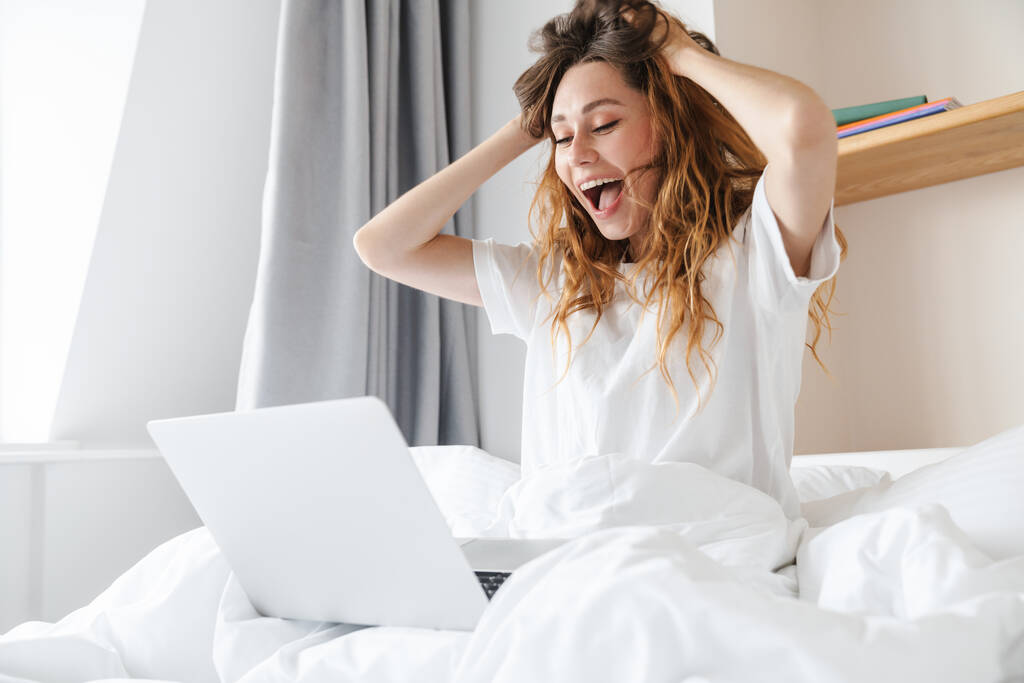 Portré gyömbéres izgatott nő kifejező meglepetés és használata laptop, miközben ül az ágyban alvás után - Fotó, kép