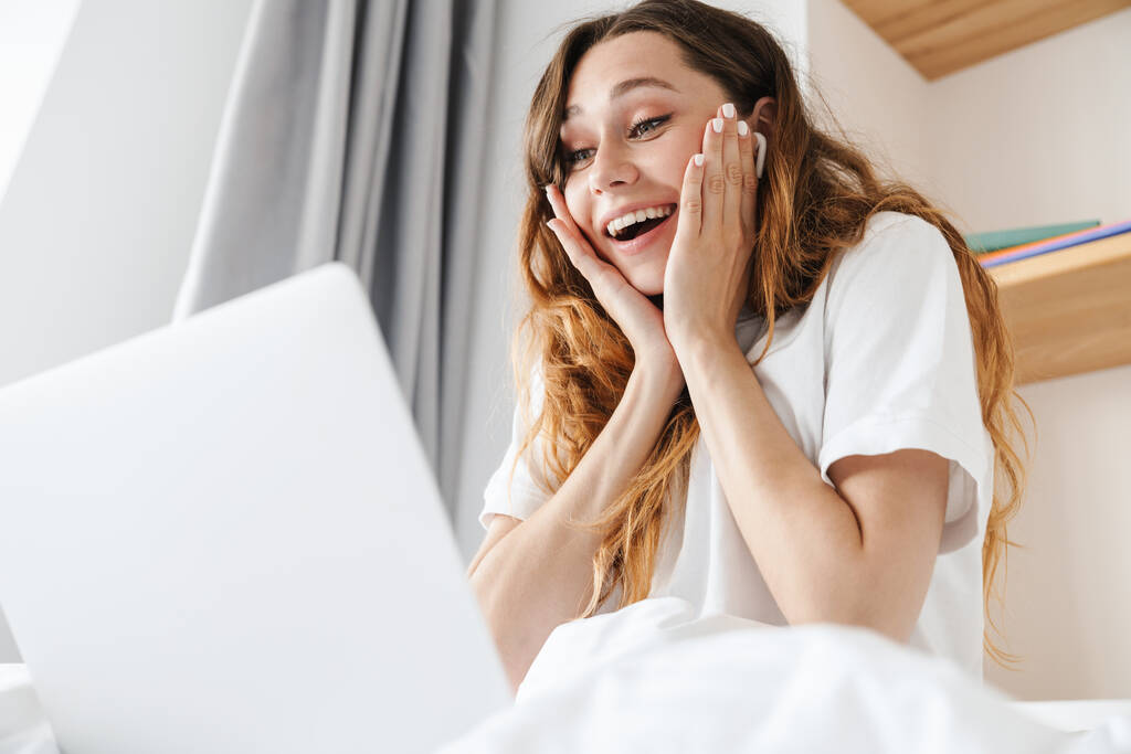 Retrato de mulher excitada com fone de ouvido expressando surpresa e sorrindo ao usar laptop na cama
 - Foto, Imagem