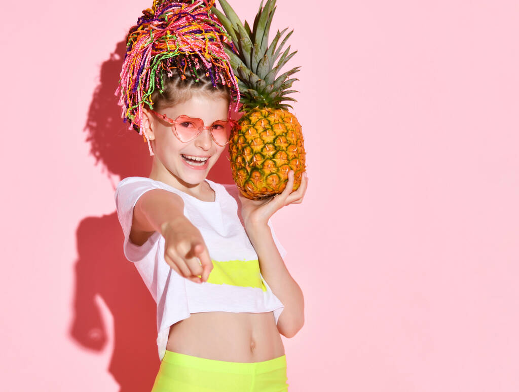 Fille active avec des tresses africaines de couleur arc-en-ciel sur la tête tenant l'ananas et pointant vers la caméra avec un sourire joyeux. Coup de feu isolé sur rose - Photo, image