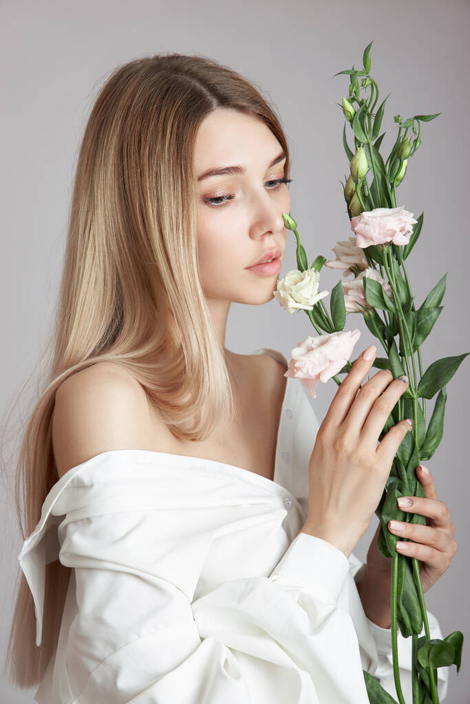 Ritratto di bellezza donna con fiori di eustoma rosa nelle sue mani. Cosmetici naturali per la cura del viso e del corpo. Bella ragazza in camicia bianca - Foto, immagini