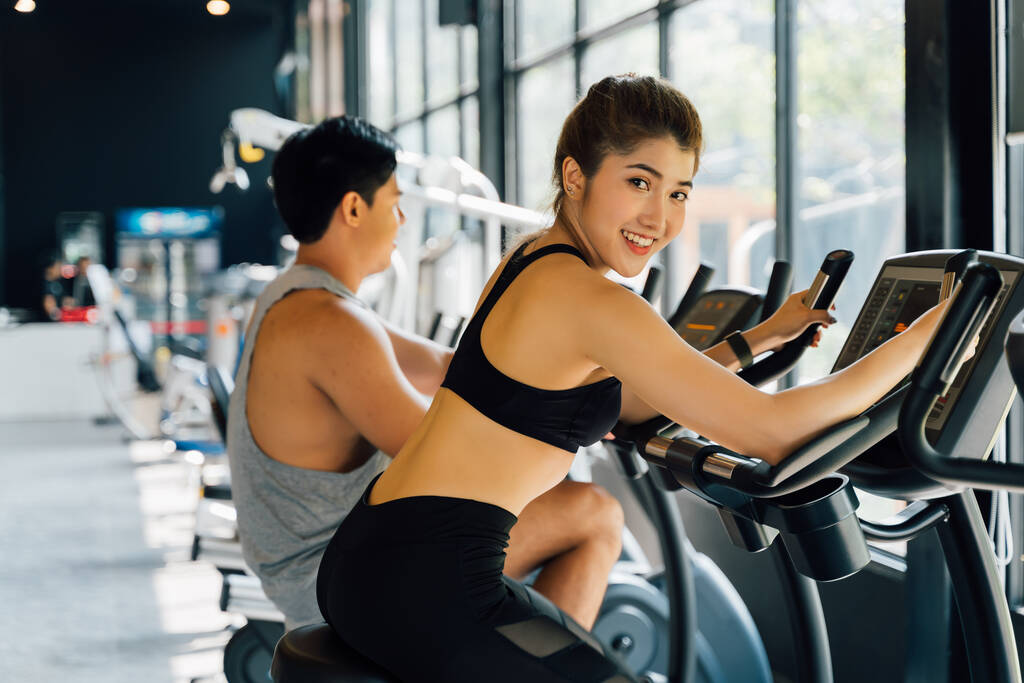 Atraktivní asijská žena jezdí na točícím se kole v tělocvičně a dívá se na kameru. Zdravý a hubnutí životní styl - Fotografie, Obrázek