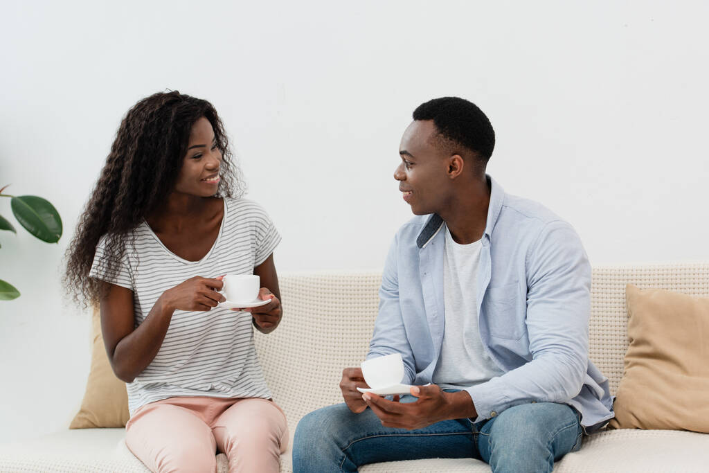 coppia afro-americana in possesso di tazze di caffè e seduto sul divano  - Foto, immagini