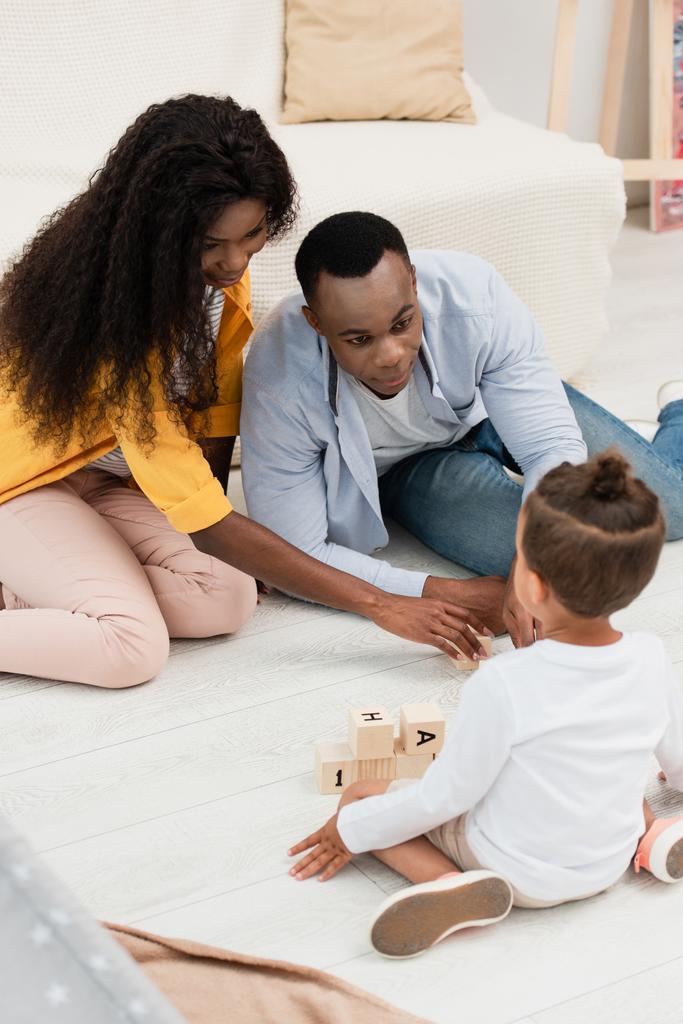 Selektiver Fokus afrikanisch-amerikanischer Eltern, die ihr Kind auf dem Boden in der Nähe von Holzwürfeln sitzen sehen  - Foto, Bild