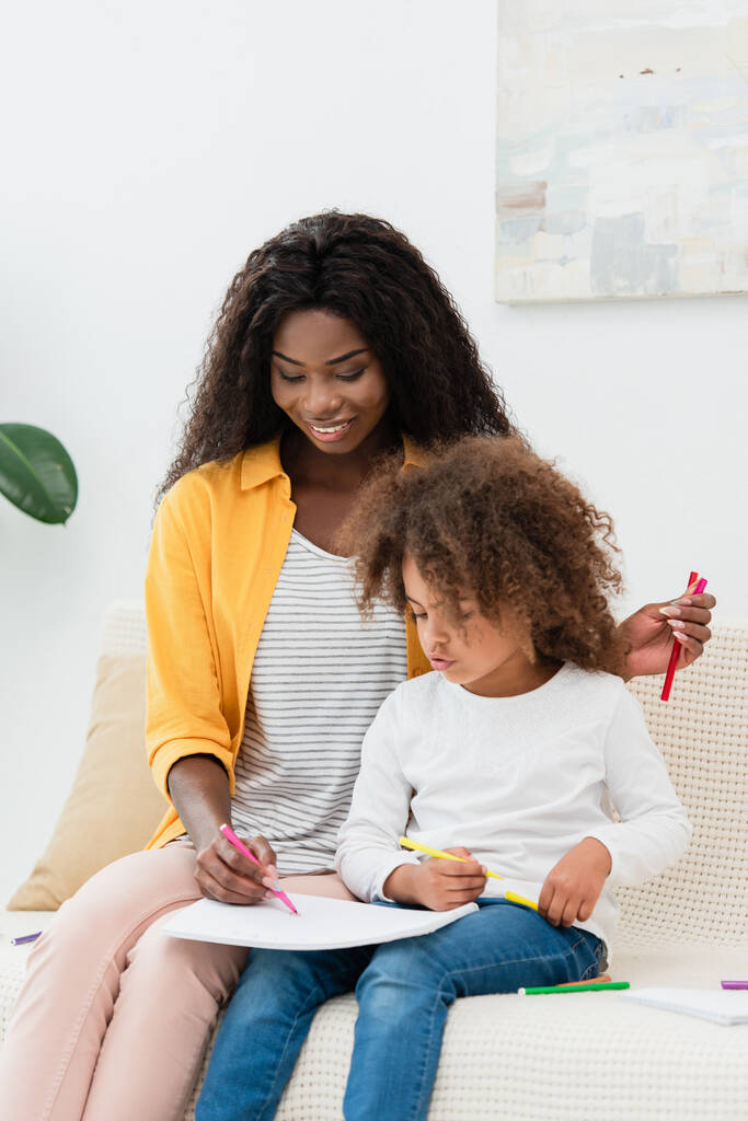 Африканская американская мать рисует цветным карандашом, сидя на диване с кудрявой дочерью  - Фото, изображение