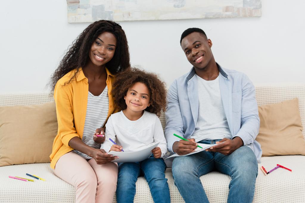 Afrikanisch-amerikanische Familie zeichnet mit Farbstiften, während sie auf dem Sofa im Wohnzimmer sitzt  - Foto, Bild