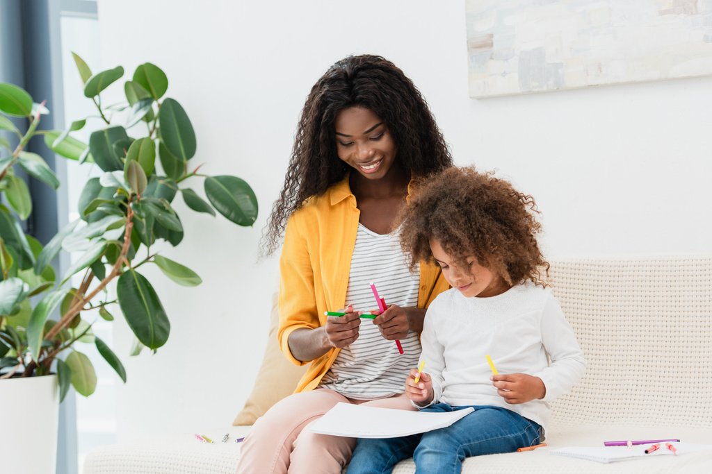 africano americano madre holding color lápiz mientras sentado en sofá con rizado hija  - Foto, Imagen
