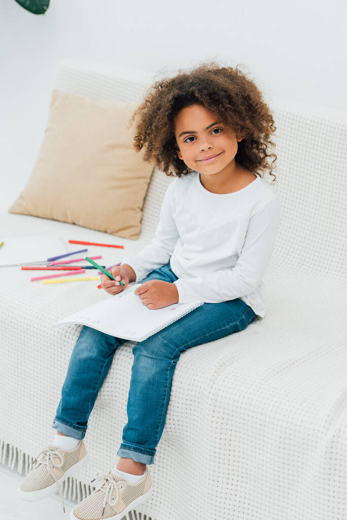 Kıvırcık Afrikalı Amerikalı çocuk elinde renkli kalemle kameraya bakıyor.  - Fotoğraf, Görsel