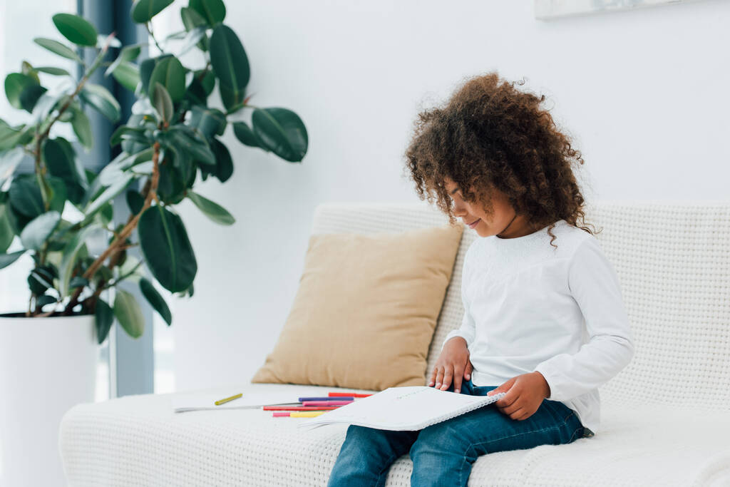 アフリカ系アメリカ人の子供が白い紙を見ながら色鉛筆の近くのソファに座り  - 写真・画像