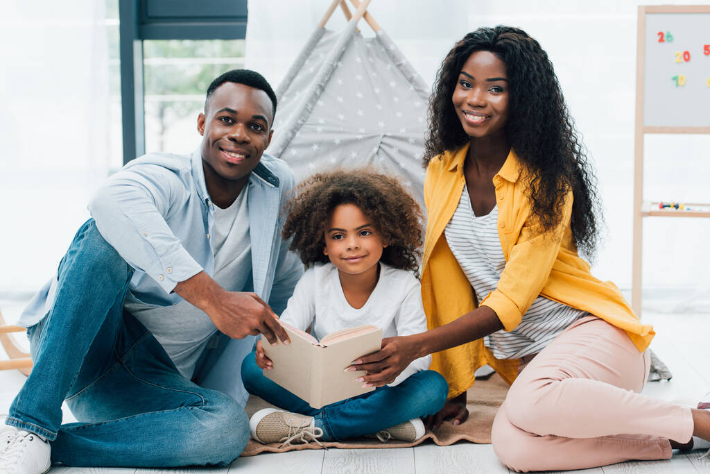 Afričtí američtí rodiče a kudrnaté dítě drží knihu, zatímco sedí na podlaze  - Fotografie, Obrázek