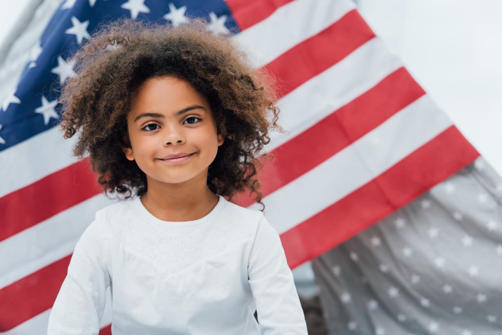 Kıvırcık Afrikalı Amerikalı çocuk Amerika bayrağının yanındaki kameraya bakıyor. - Fotoğraf, Görsel
