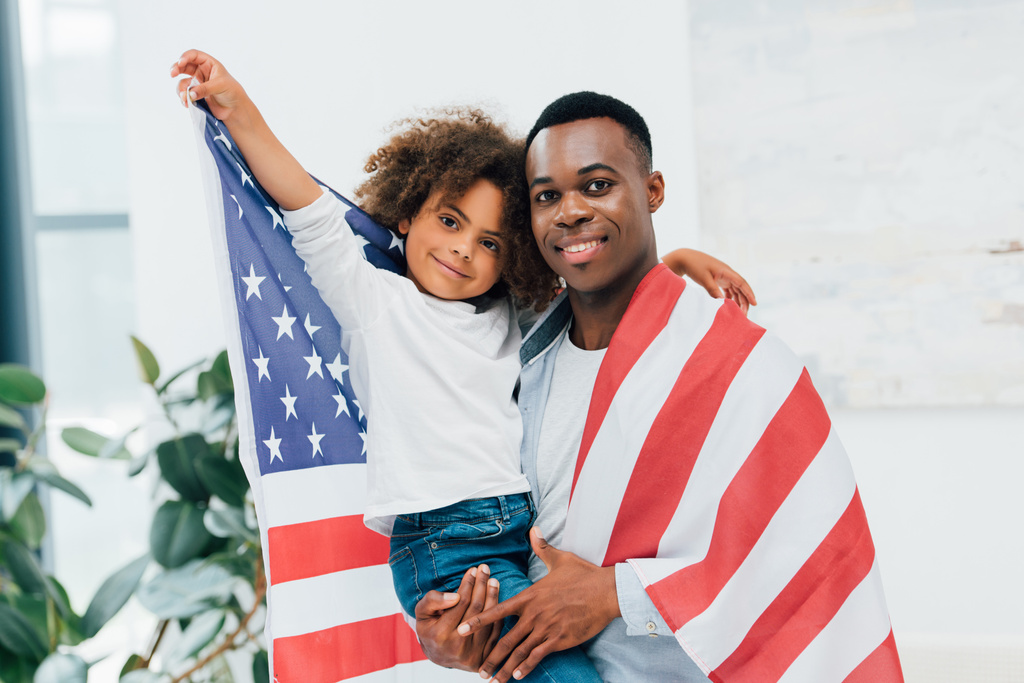 Αφροαμερικάνος πατέρας και κόρη τυλιγμένα με σημαία Αμερικής κοιτώντας την κάμερα  - Φωτογραφία, εικόνα