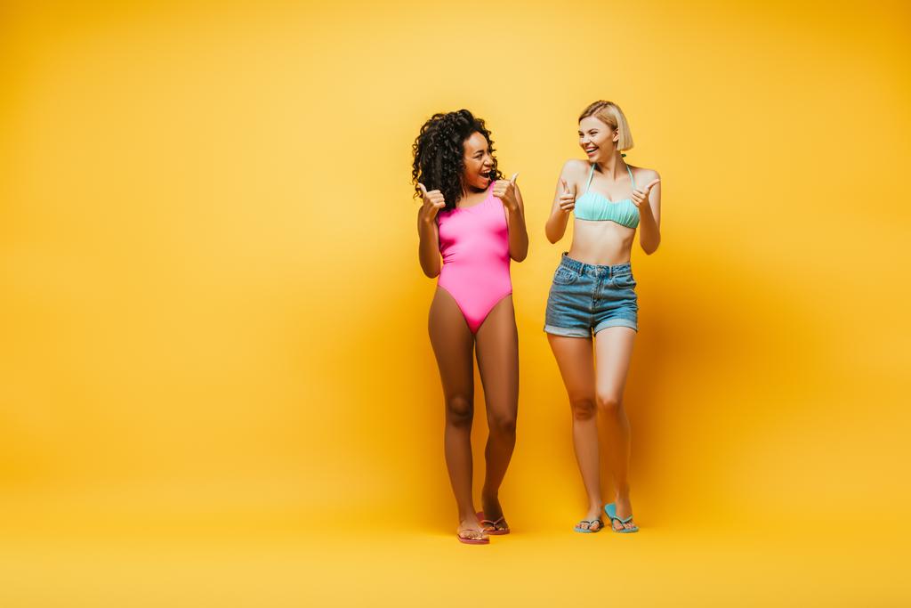 full length view of excited interracial women showing thumbs up and looking at each other on yellow - Photo, Image