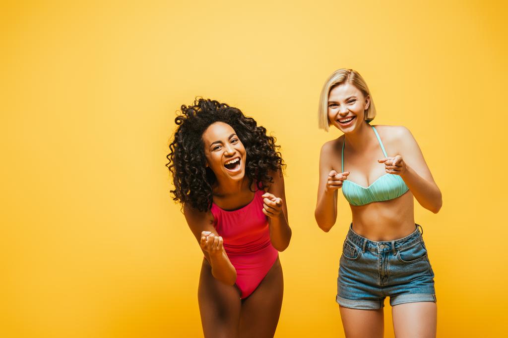 興奮した異人種間の女性の夏の衣装は笑いと黄色に隔離されたカメラで指を指して - 写真・画像