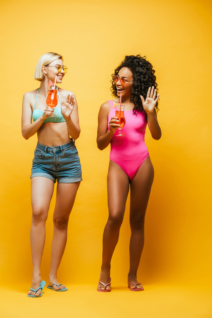 joven afroamericana americana mujer mostrando hola gesto cerca rubia amigo mientras sosteniendo copas de cóctel en amarillo - Foto, imagen