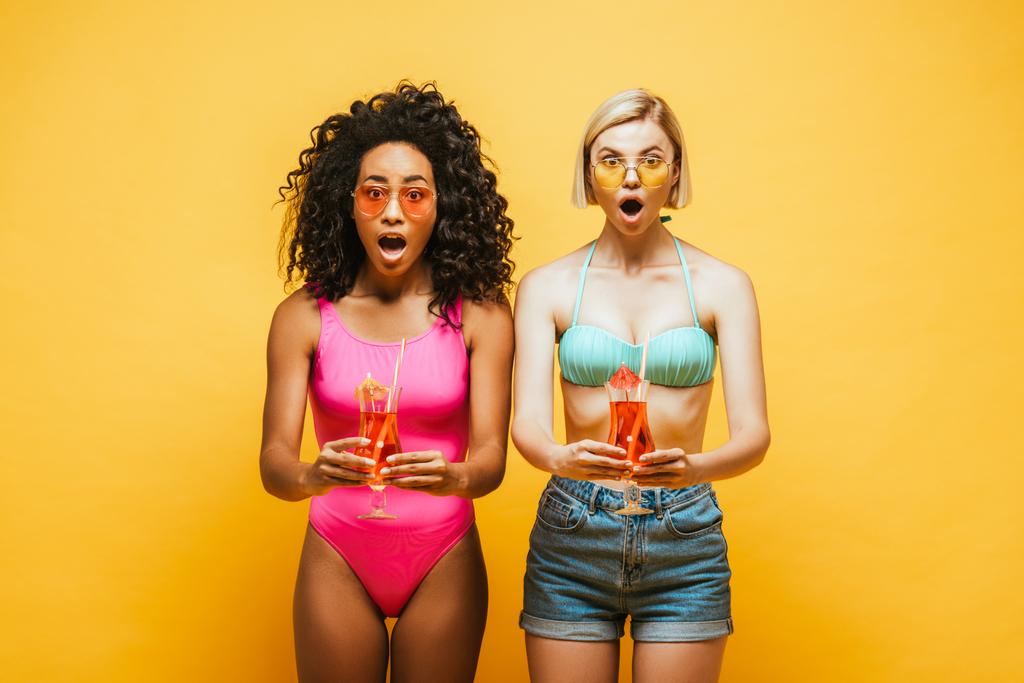 excitado interracial mujeres en verano traje celebración copas de cóctel y mirando a la cámara en amarillo - Foto, Imagen