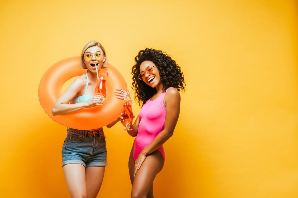 excitées femmes interracial avec anneau de natation et verres à cocktail posant sur jaune - Photo, image