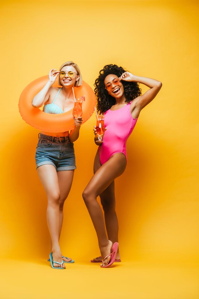 возбужденные межрасовые друзья с коктейлями и кольцами для купания касательно солнцезащитных очков на желтом - Фото, изображение