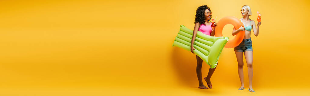 imagen horizontal de mujeres jóvenes multiétnicas en traje de verano posando con colchón inflable, anillo de natación y cócteles en amarillo - Foto, imagen