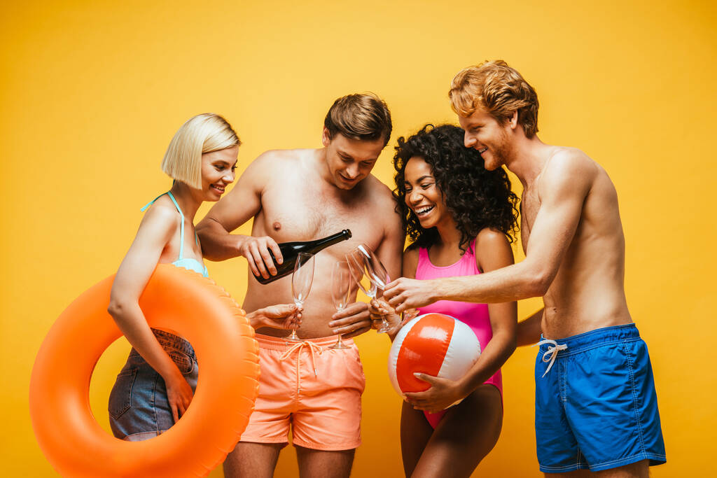 shirtless man pouring champagne in glasses of multiethnic friends with inflatable ball and ring isolated on yellow - Photo, Image