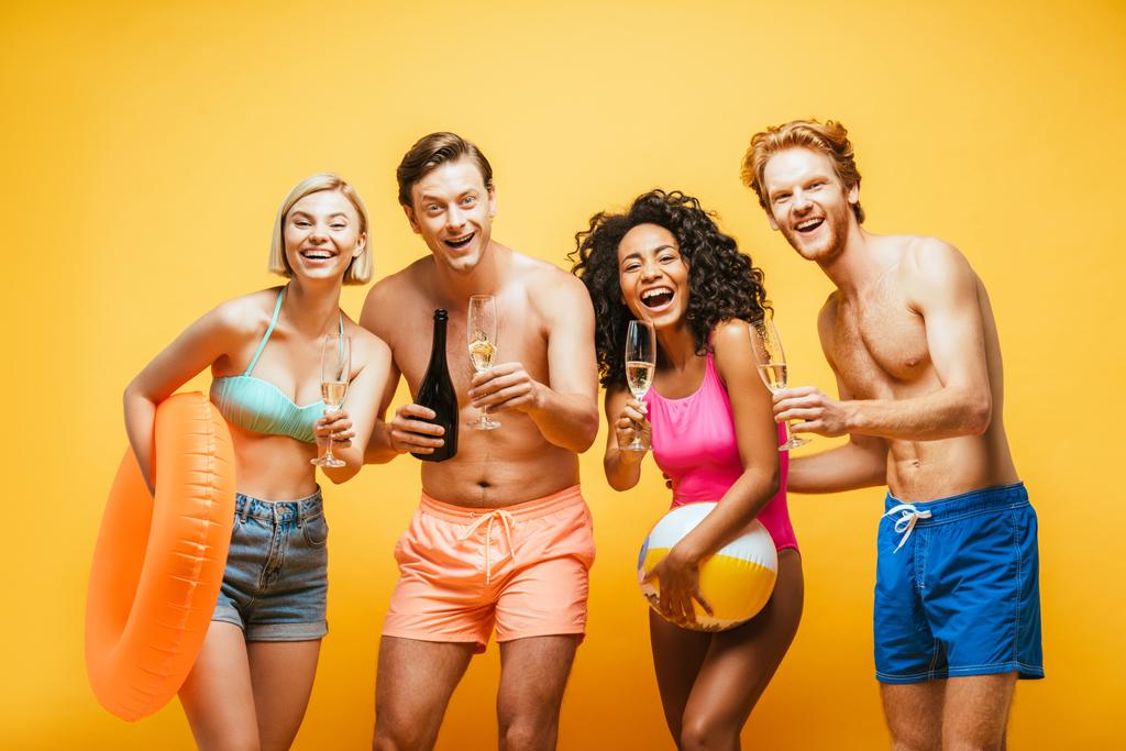 возбужденные мультикультурные друзья с бокалами шампанского, надувные кольца и мяч глядя на камеру изолированы на желтый - Фото, изображение