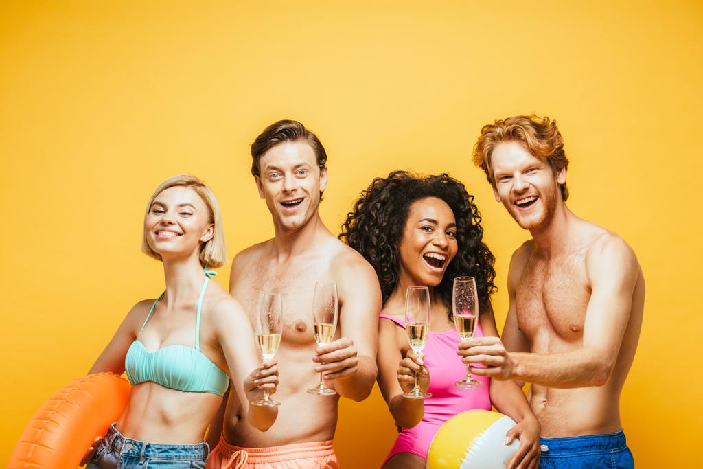 aufgeregte multikulturelle Freunde blicken in die Kamera, während sie Champagnergläser isoliert auf gelb halten - Foto, Bild