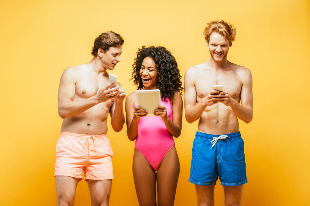 amigos multiculturais excitados em roupas de verão usando dispositivos digitais isolados em amarelo
 - Foto, Imagem