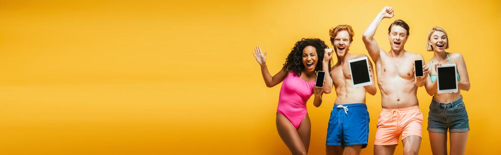 горизонтальное изображение взволнованных мультикультурных друзей в летнем наряде, показывающих гаджеты с чистым экраном на желтом - Фото, изображение